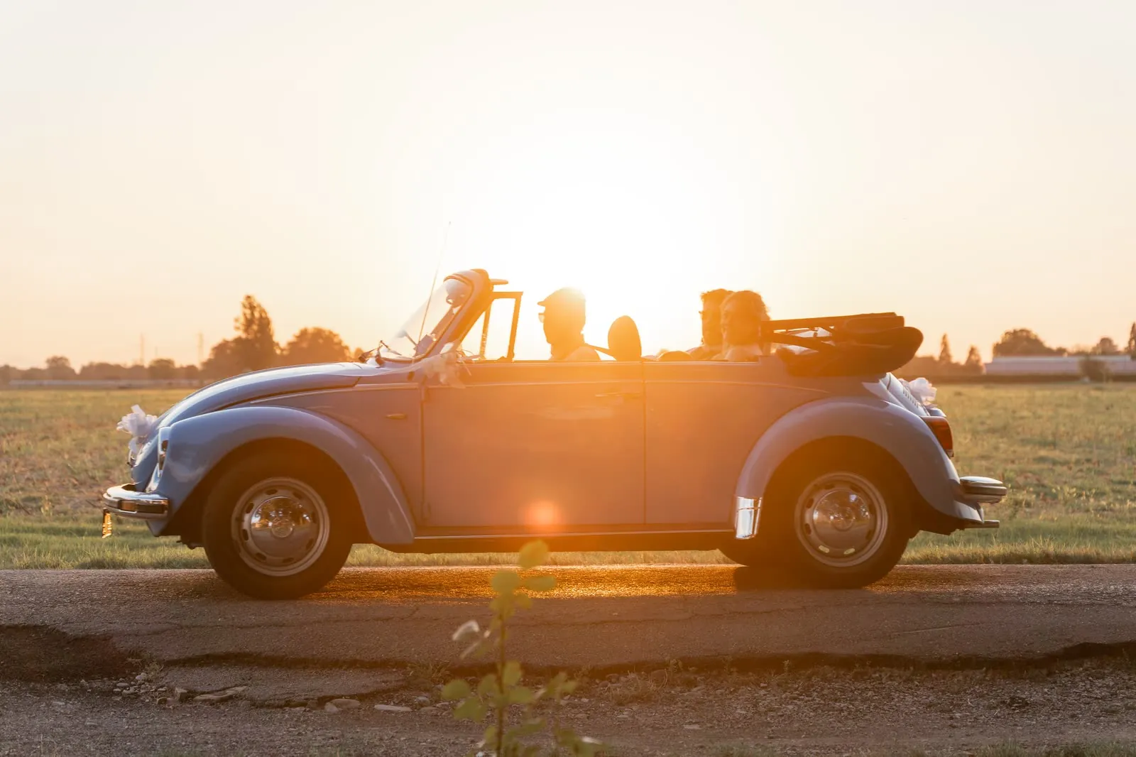 Il Maggiolone cabriolet azzurro degli sposi in controluce su una strada di campagna al tramonto, con il sole alle spalle e i fiocchi bianchi sui parafanghi