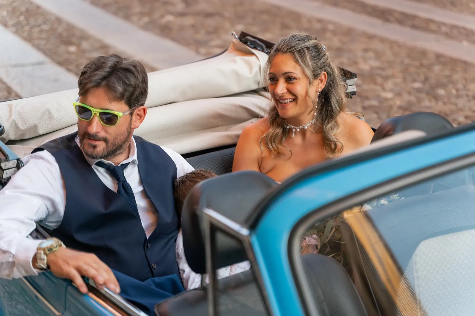 Gli sposi seduti nel Maggiolone cabriolet azzurro con la capote abbassata, in piazza: lui con gli occhiali da sole giallo fluo, lei che sorride