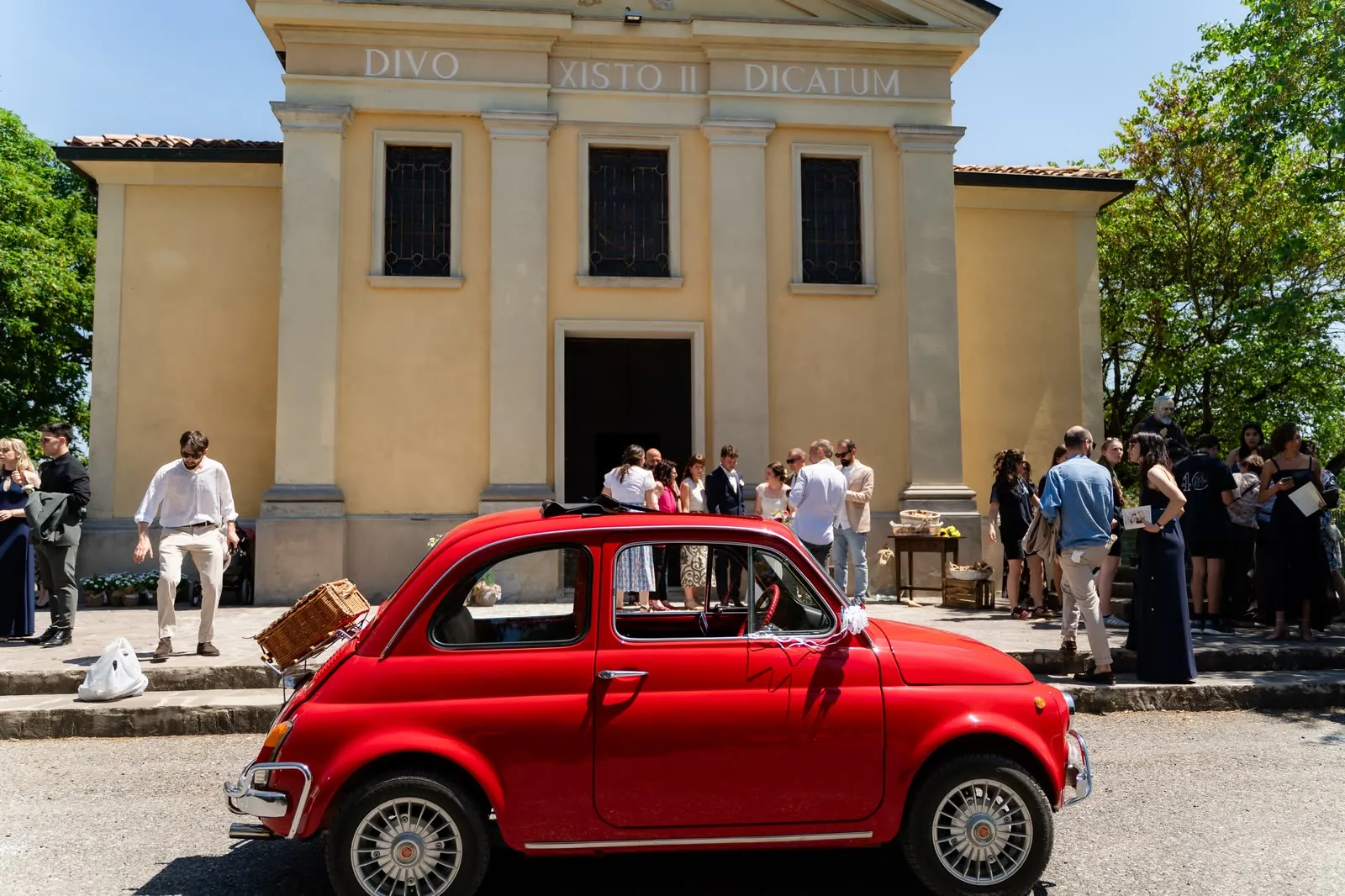 La Fiat 500 rossa d'epoca degli sposi davanti alla chiesetta gialla, con gli invitati sul sagrato
