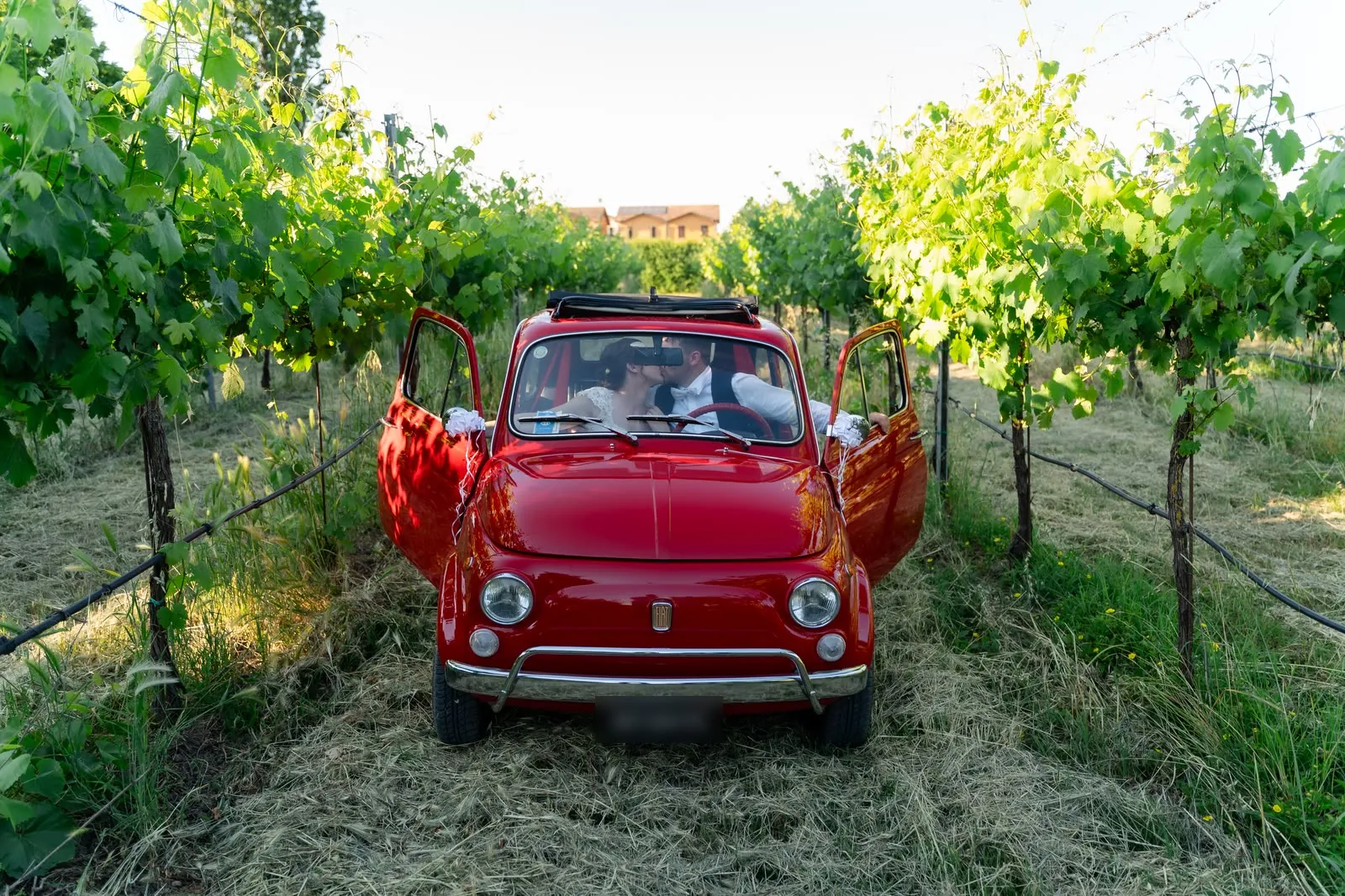 La Fiat 500 rossa d'epoca tra i filari della vigna di casa, con le portiere aperte e gli sposi che si baciano all'interno; in fondo la casa