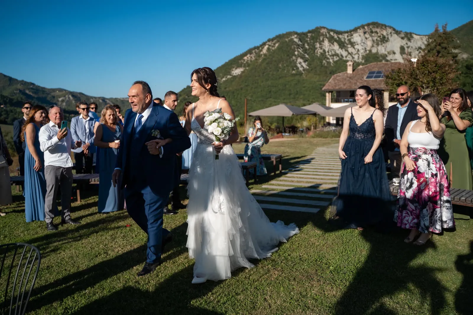 La sposa arriva sul prato a braccio di un uomo in abito blu, sorridente, mentre gli invitati la guardano e la fotografano